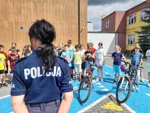 Policjantka ustawiona tyłem, w tle dzieci odbierające karty rowerowe