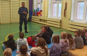 na zdjęciach widoczna jest szkolna sala gimnastyczna a w niej umundurowany policjant, który stoi przed dziećmi. Policjant ma ze sobą kamizelki odblaskowe.
