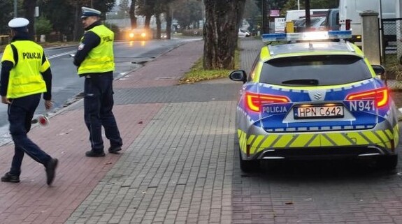 Na zdjęciu widoczna jest droga, oznakowany radiowóz i dwóch umundurowanych policjantów.
